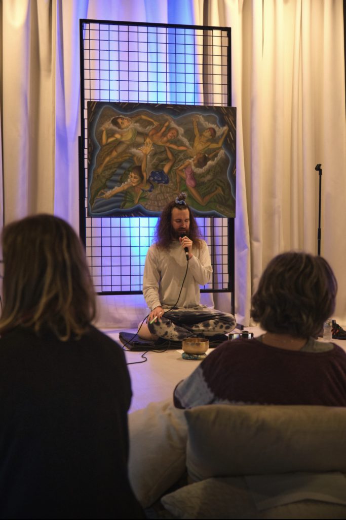 David Prorok seated cross-legged, leading a meditation session with a microphone in one hand and a marker in the other, in front of an audience and a vibrant spiritual painting